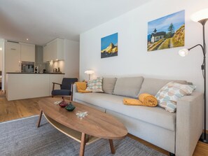 Ein modernes Wohnzimmer mit Sofa, Couchtisch, Stuhl und einer Küche im Hintergrund.