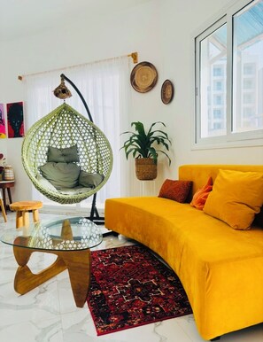A modern living room with a hanging chair, a yellow sofa, a glass coffee table, and a patterned rug. - Almadies Luxurious 2bed apartment with Ocean view (Dakar)