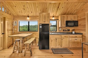 Eine Küchenzeile im Blockhausstil mit Holzwänden, einem schwarzen Kühlschrank, einem Holztisch und einem Fenster mit Blick auf Grünflächen.