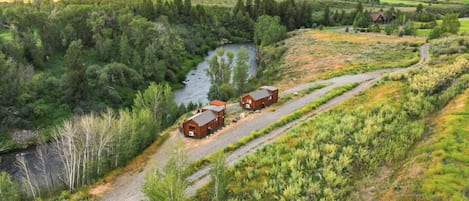 A rural landscape with a river, forested areas, and two cabins on a dirt road.