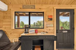 Ein Holzblockhaus-Interieur mit einem 'WELCOME'-Schild, einer Kaffeestation, einem 'NO SMOKING'-Schild und Blick ins Freie.