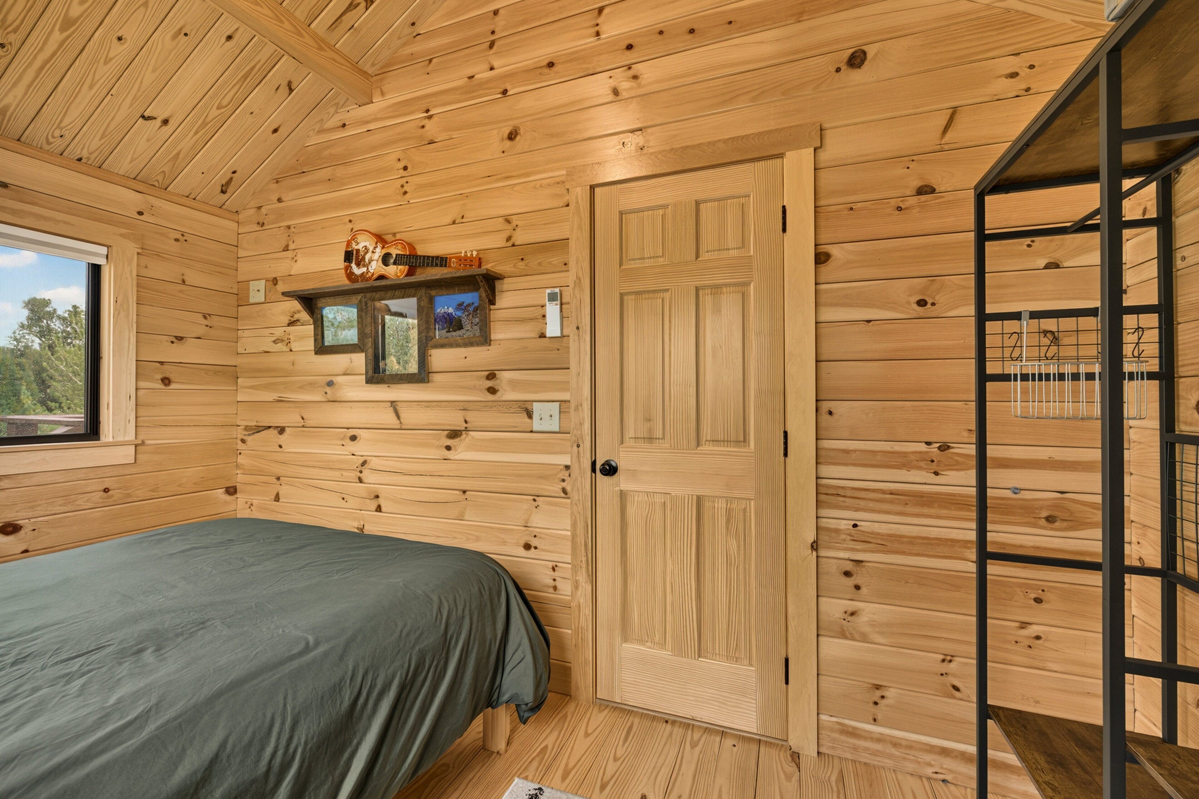 A wooden cabin room with a bed, a door, a shelf with pictures, and a window.