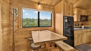 Interior d'una cabana de fusta amb una taula de dinar, un frigorífic i una finestra amb vista a vegetació.