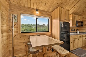A wooden cabin interior with a dining table, a refrigerator, and a window overlooking greenery. - 2BR cabin near water, mountains, and outdoor adventure. (Ashton)