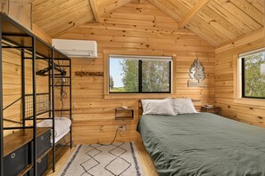 A wooden cabin bedroom with bunk beds, a bed, a window, and a wall-mounted air conditioner. - 2BR cabin near water, mountains, and outdoor adventure. (Ashton)