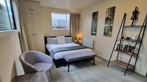 Habitación de hotel pequeña con cama, dos sillones, una mesita, ventana con cortinas, radiador y estante de pared con objetos decorativos.