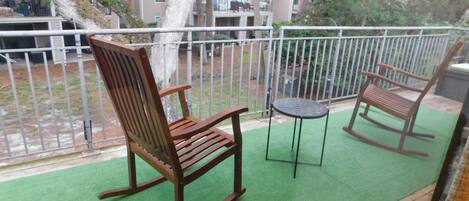 A balcony with a green artificial turf, a wooden rocking chair, a small round table, and a metal chair.