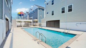 Uma piscina com deck, área coberta e um edifício com várias janelas e porta de garagem.