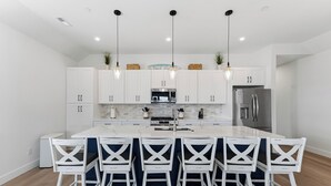 Uma cozinha moderna com armários brancos, uma ilha central com banquetas, eletrodomésticos de aço inoxidável e iluminação pendente.