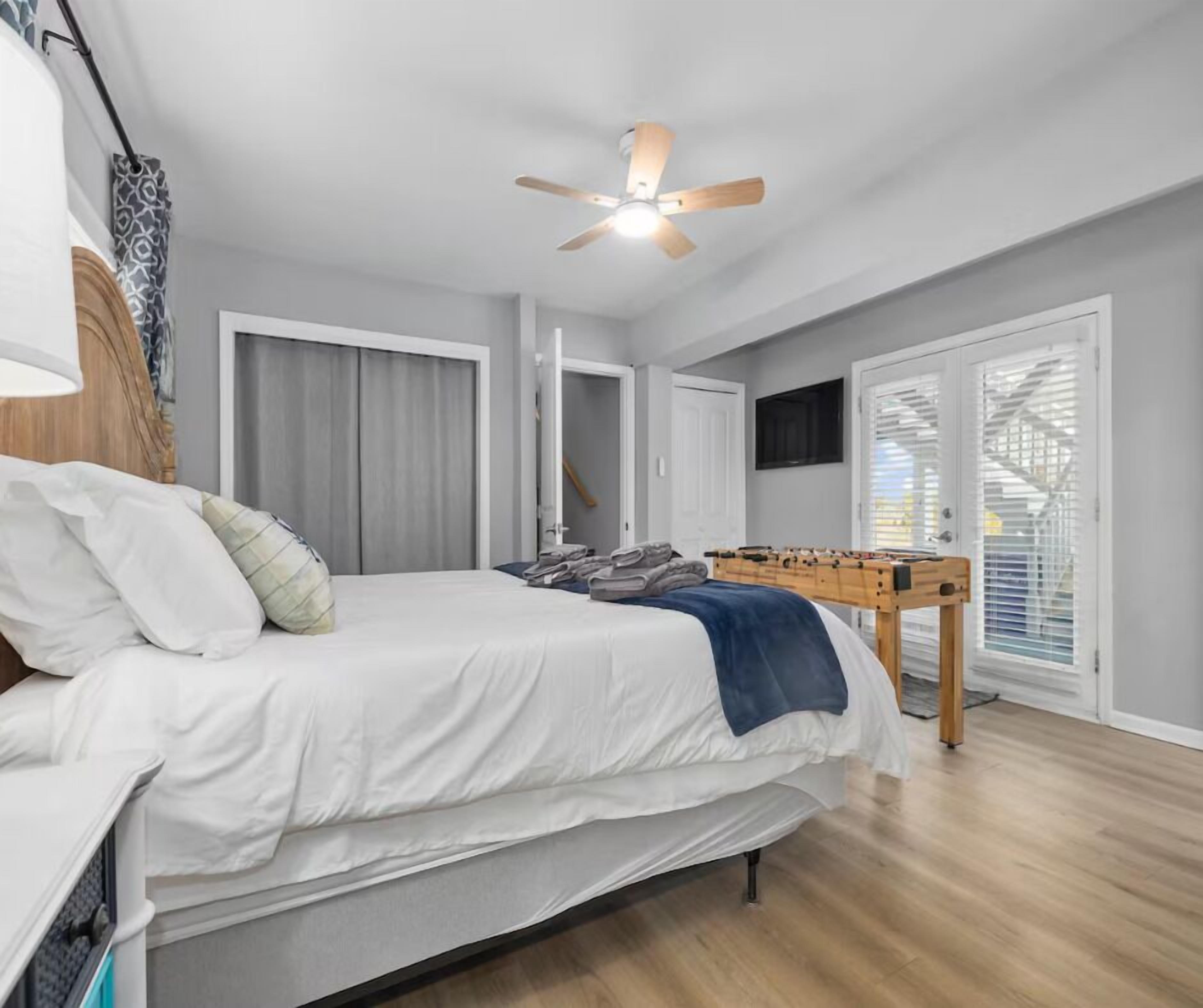 A bedroom with a bed, a ceiling fan, a window with blinds, and a foosball table.