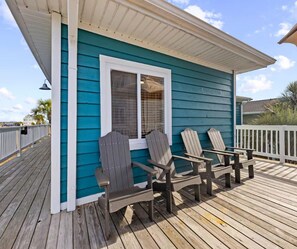 Eine Holzterrasse mit Adirondack-Stühlen, einem weißen Geländer und einem türkisblauen Gebäude mit einem Fenster.