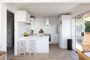 A modern kitchen with white cabinetry, a central island with stools, a built-in oven, and a large window. - Entire home in Waimārama, sea views, 2 minute walk to the beach. (Waimārama)