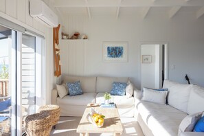 A living room with a sofa, a coffee table with fruit, a wicker chair, and a guitar in the background. - Entire home in Waimārama, sea views, 2 minute walk to the beach. (Waimārama)