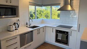 A modern kitchen with white cabinetry, a built-in microwave, a stove, and a window overlooking greenery. - Spacious Waimārama Beach House with Large Deck (Waimārama)