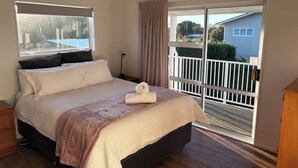 A bedroom with a bed, a nightstand, a window with blinds, a balcony with a railing, and a view of the outside. - Spacious Waimārama Beach House with Large Deck (Waimārama)