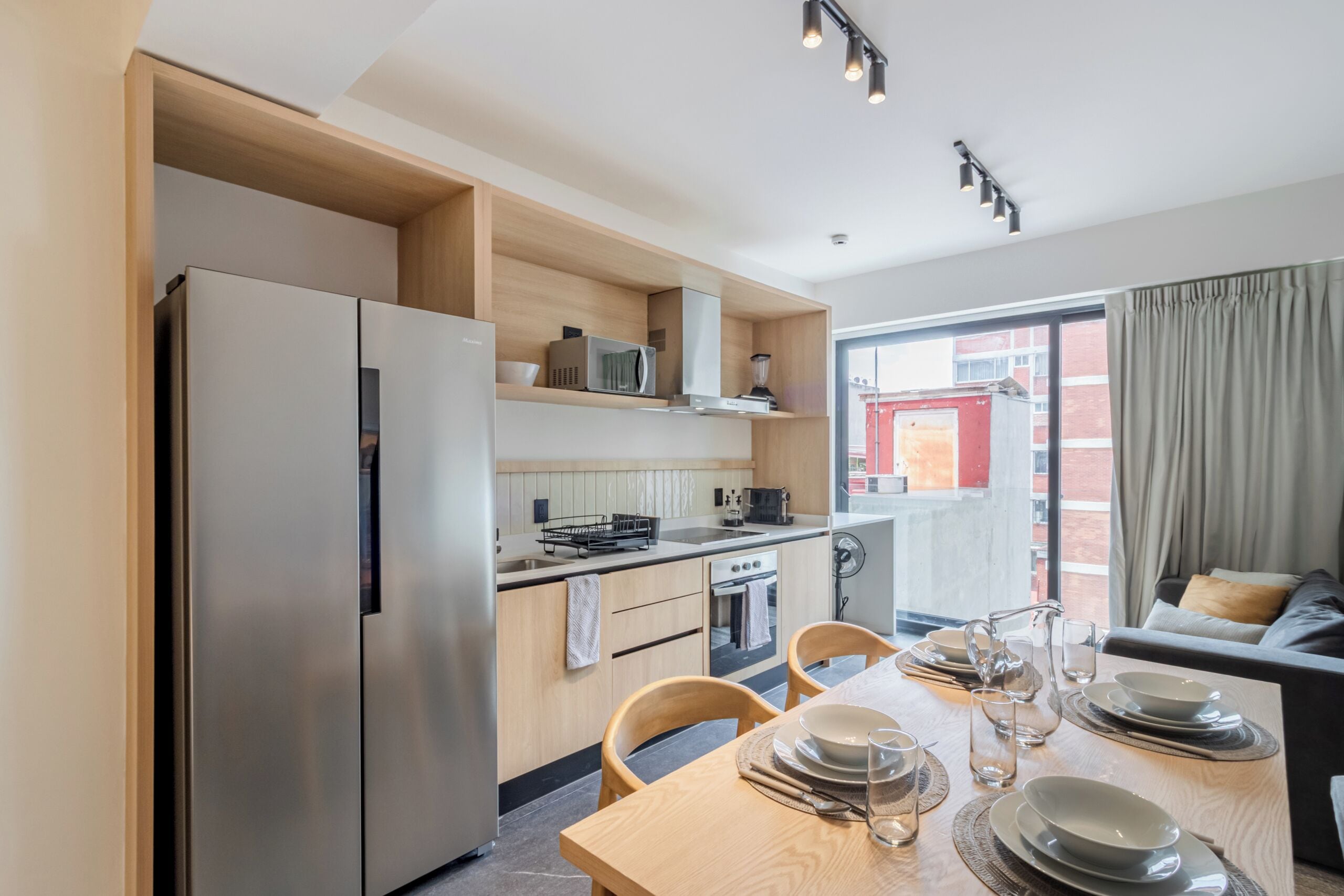 A modern kitchen with a refrigerator, microwave, and stove, a dining table set with plates and glasses, and a view of a red building through a window.