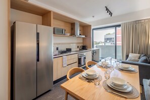 Eine moderne Küche mit Kühlschrank, Mikrowelle und Herd. Ein Esstisch ist mit Tellern, Gläsern und Besteck gedeckt. Im Hintergrund sind eine Couch und ein Fenster mit Vorhängen zu sehen.