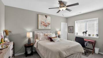 Un dormitorio con cama, lámparas de noche, escritorio, silla, ventilador de techo y ventana con persianas.