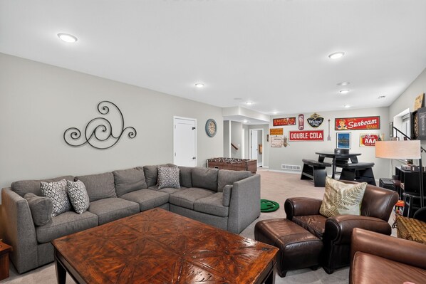 A modern living room with a sectional sofa, a coffee table, and a leather ottoman. There are wall decorations including a metal wall art piece and vintage-style signs.