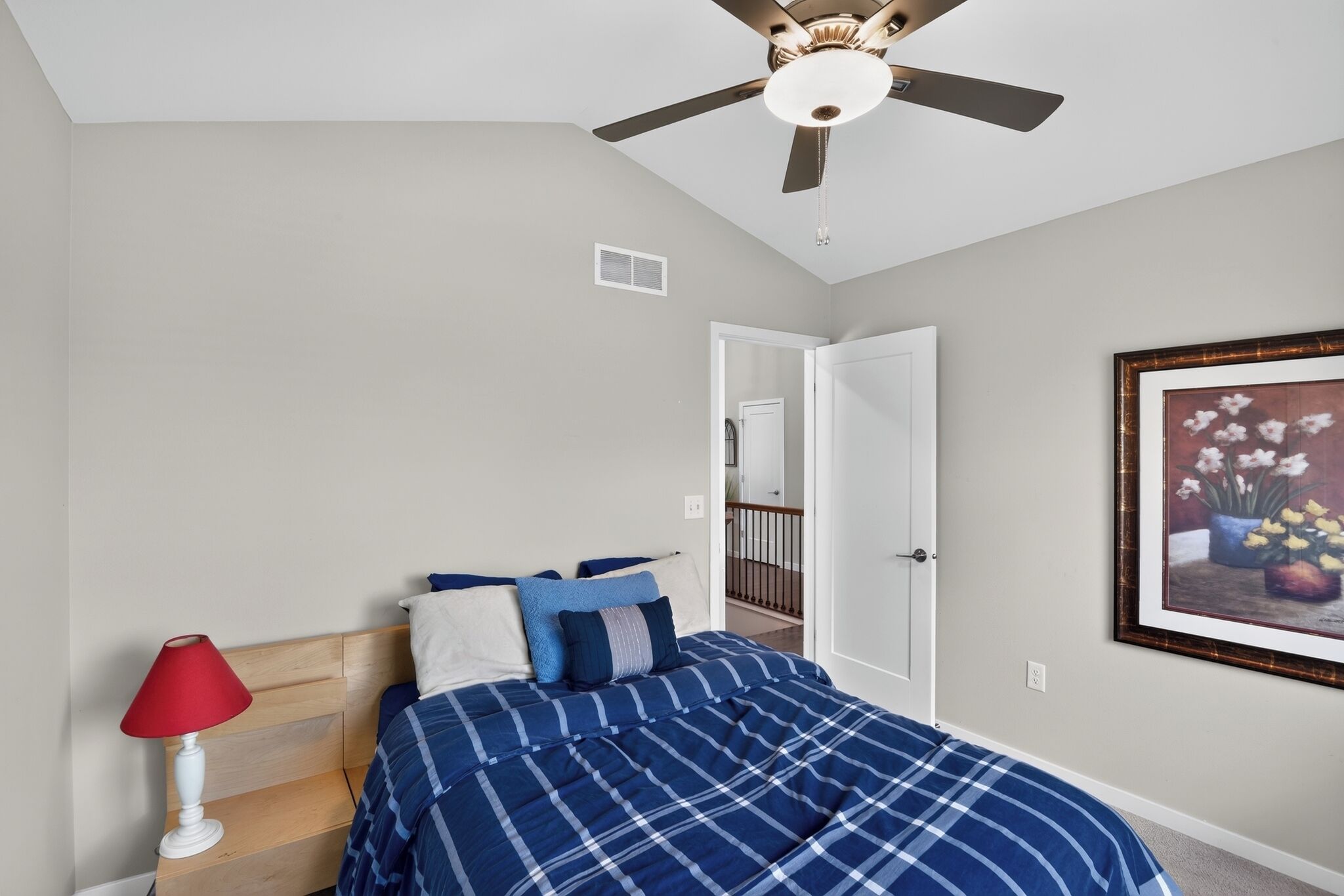 A bedroom with a bed, a ceiling fan, a red lamp, and a framed picture on the wall.