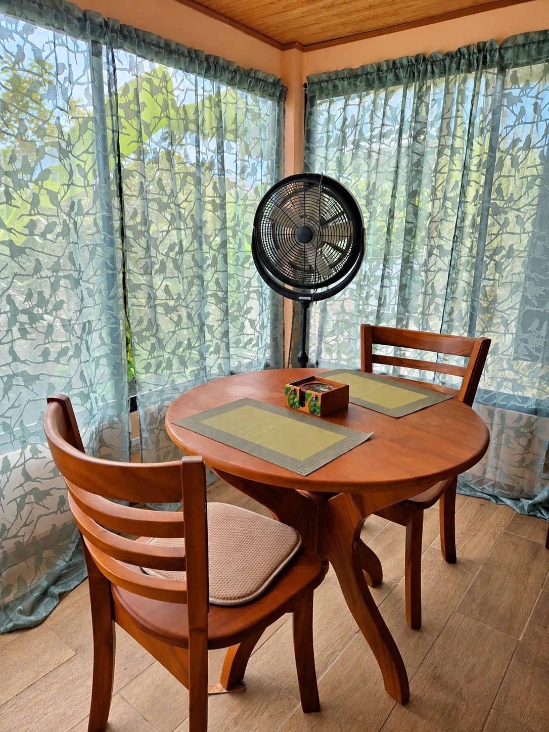 A dining area with a round wooden table, two chairs, a fan, and patterned curtains.