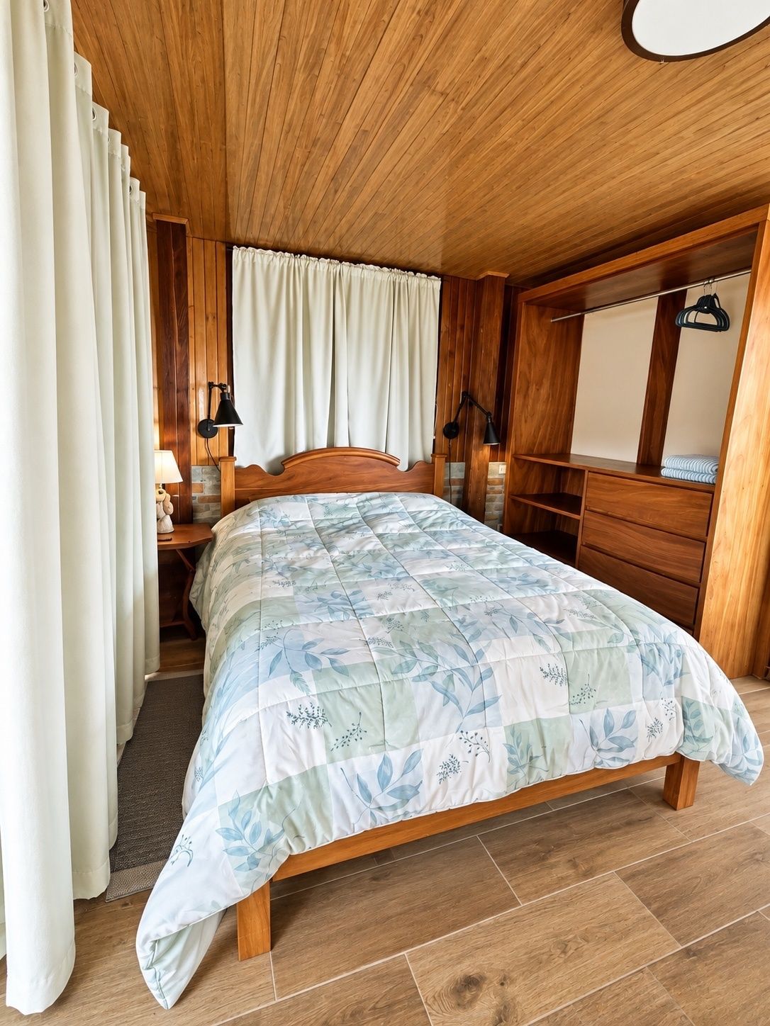 Une chambre avec un plafond en bois, un lit recouvert d’une couvre-lit à motifs floraux, une table de chevet avec une lampe et une armoire en bois.