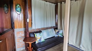 A room with a wooden ceiling, a brick wall, a black sofa with patterned cushions, a small wooden table, and a window with curtains. - Escazu Studio (San Antonio)
