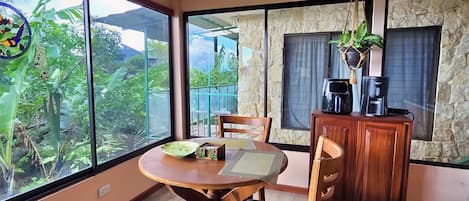 A dining area with a wooden table and chairs, a coffee station, and a view through large windows.