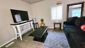 A living room with a flat-screen TV on a white stand, a dark-colored sofa, a small wooden table, and a window with blinds.