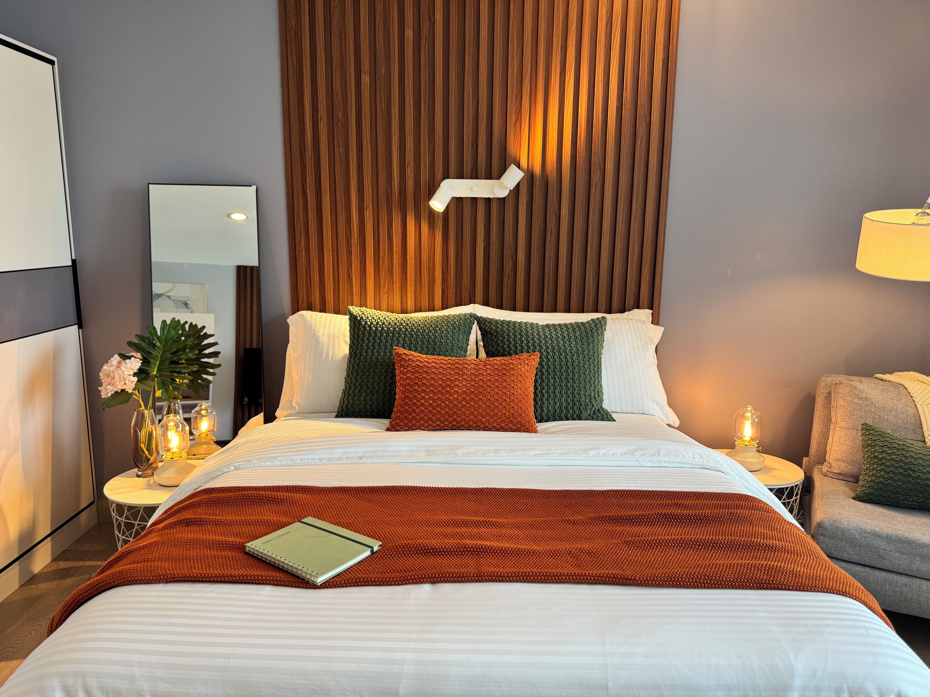 A neatly made bed with a green and orange blanket, a bedside table with a lamp, and a wall-mounted light fixture.