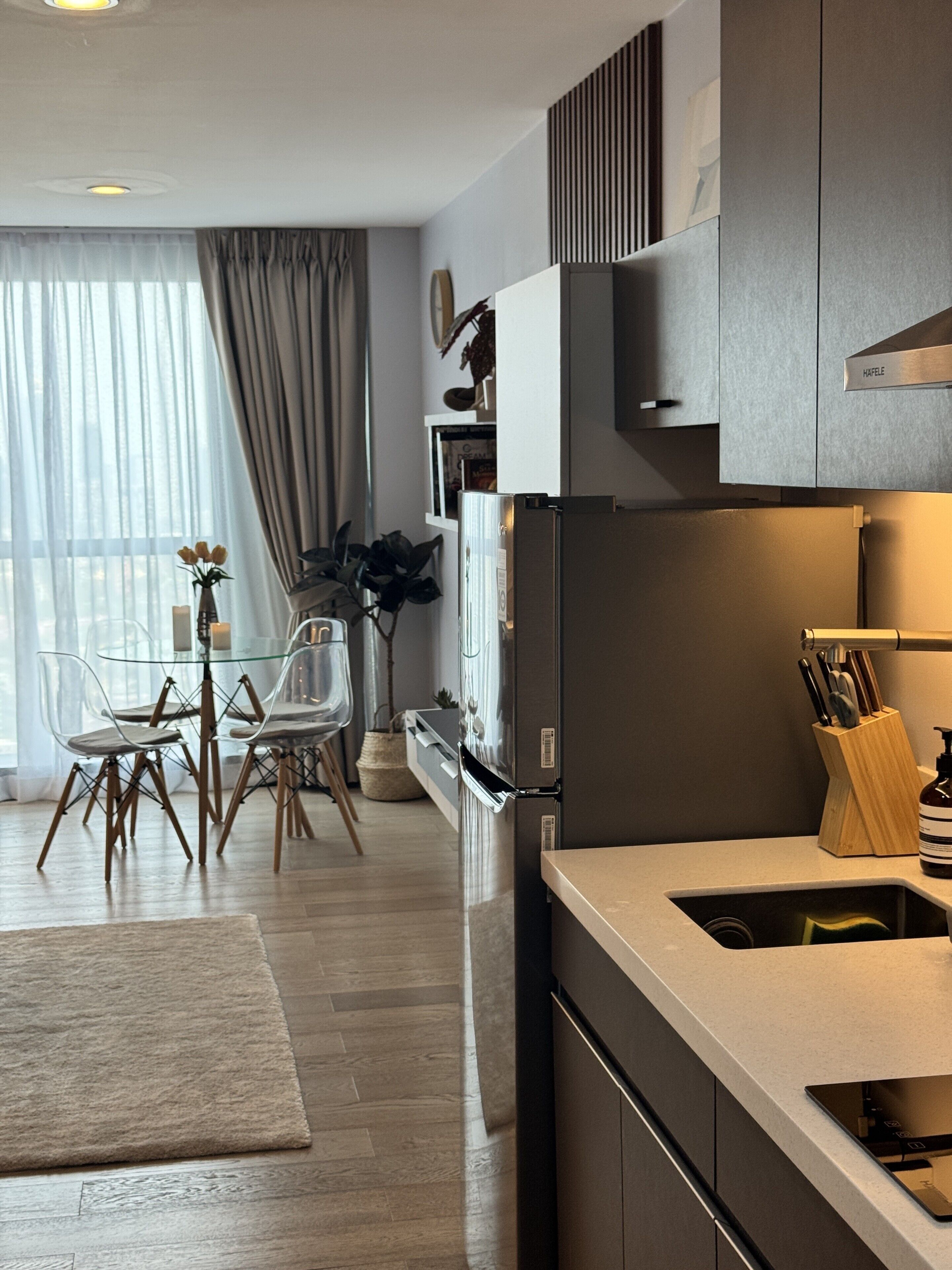 A modern kitchen with a built-in refrigerator, a dining area with a glass table and chairs, and a large window with curtains.