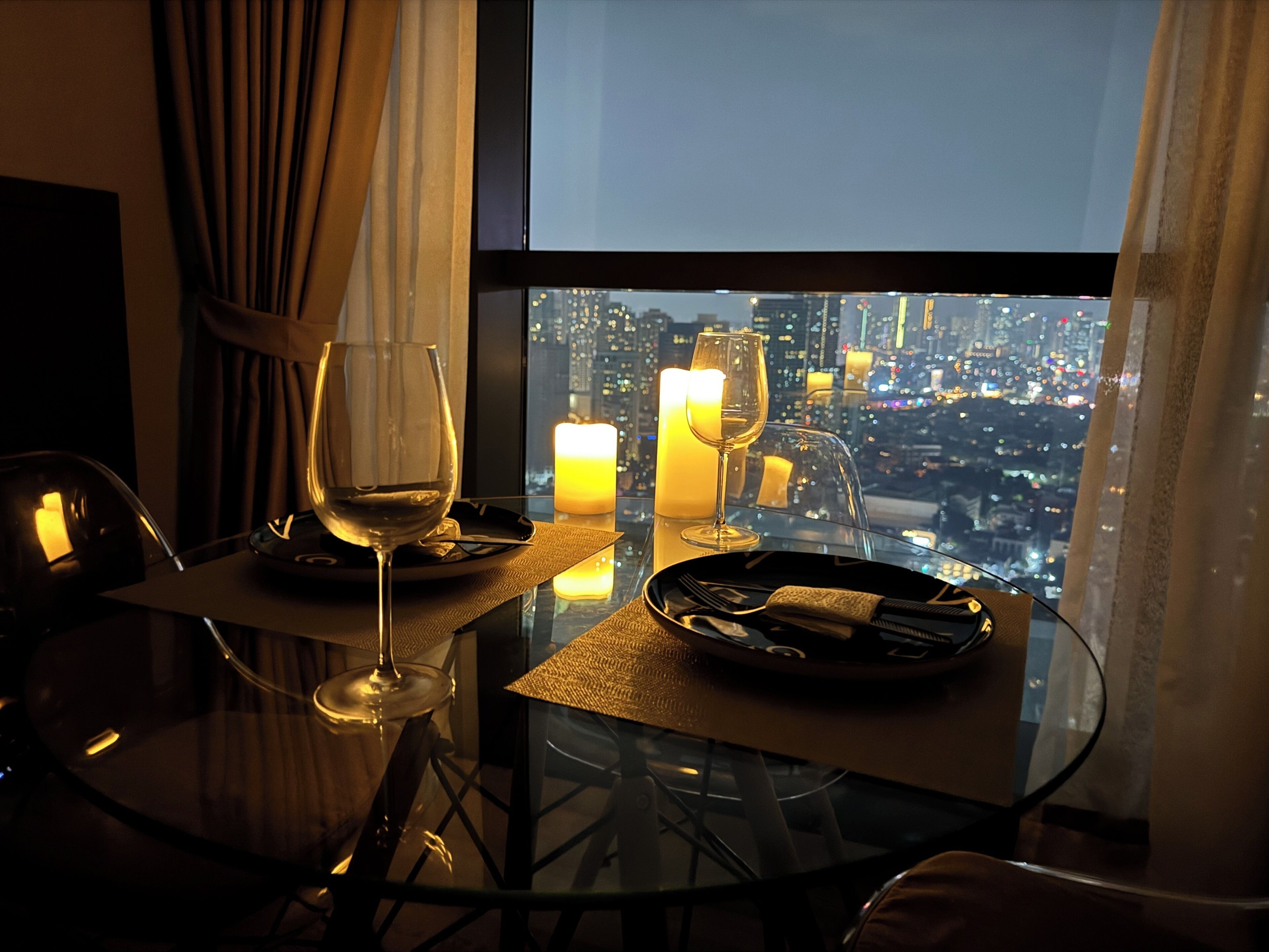 A dining table with a wine glass, candles, and a view of a cityscape at night.