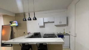 A kitchen with a marble countertop, two black bar stools, a gas stove, a microwave, and a TV mounted on the wall.
