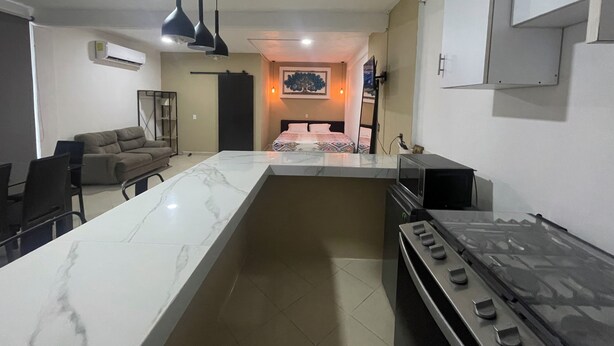 A modern kitchen with a central island, a dining area with chairs, a sofa, and a bed with a canopy in the background.