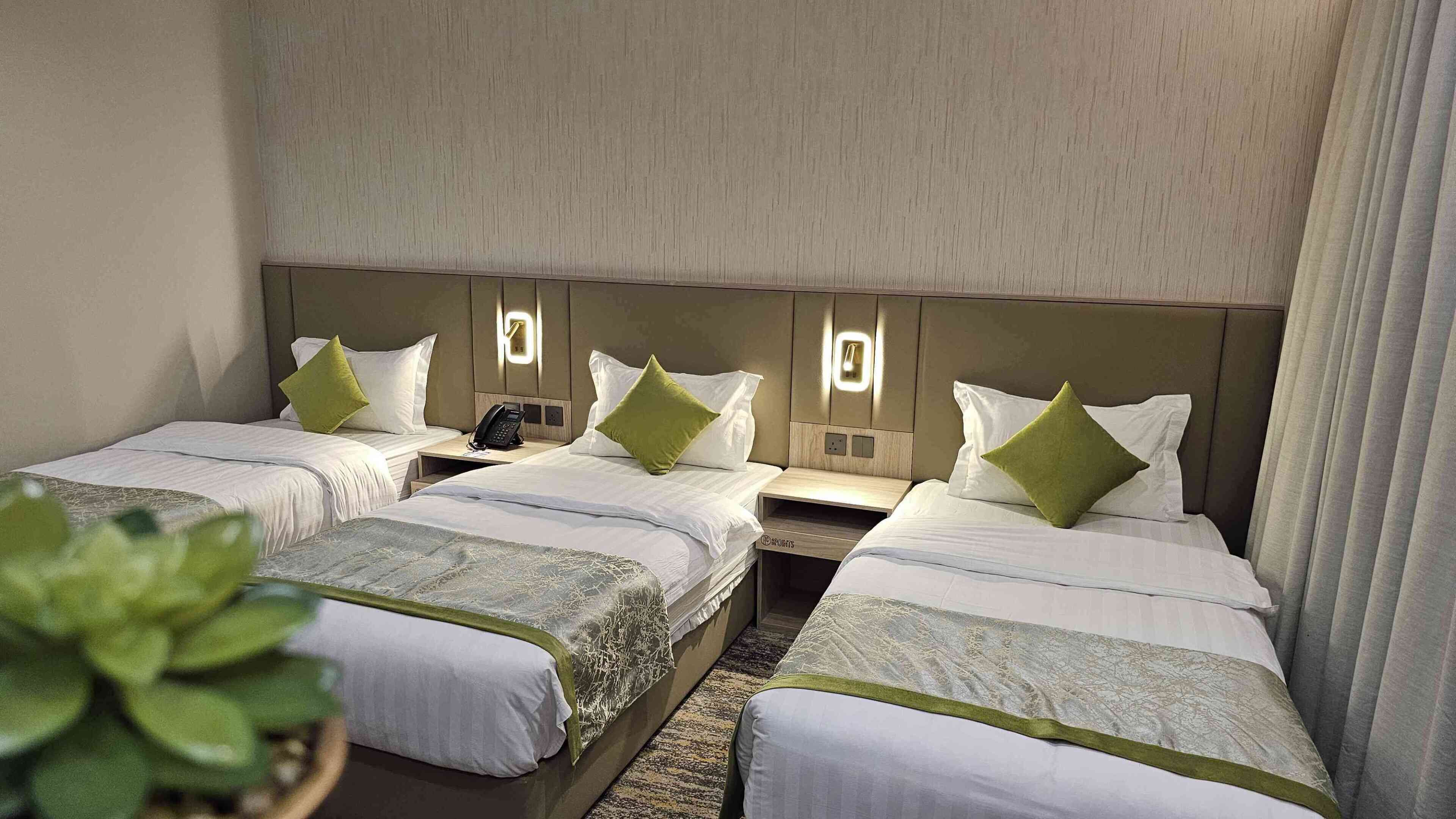 Habitación de hotel con tres camas individuales, cada una con mesita de noche y teléfono. La habitación presenta una paleta de colores neutros, con paredes color beige y una alfombra estampada.