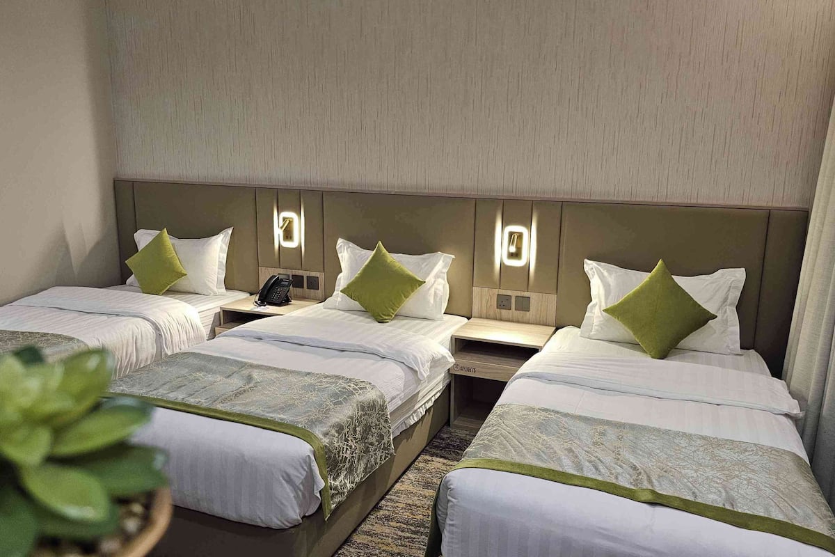 Ein Hotelzimmer mit drei Einzelbetten, jeweils mit Nachttisch und Telefon. Das Zimmer hat eine neutrale Farbgestaltung mit beigen Wänden und einem gemusterten Teppich.