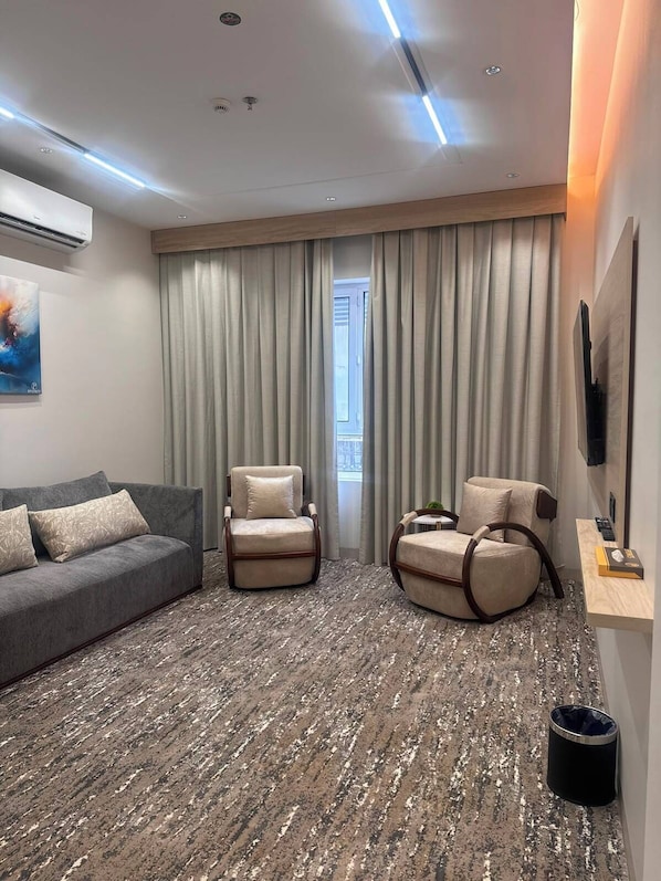 Ett modernt hotellrum med en grå soffa, två fåtöljer, en mönstrad matta, ett stort fönster med gardiner och en väggmonterad TV.