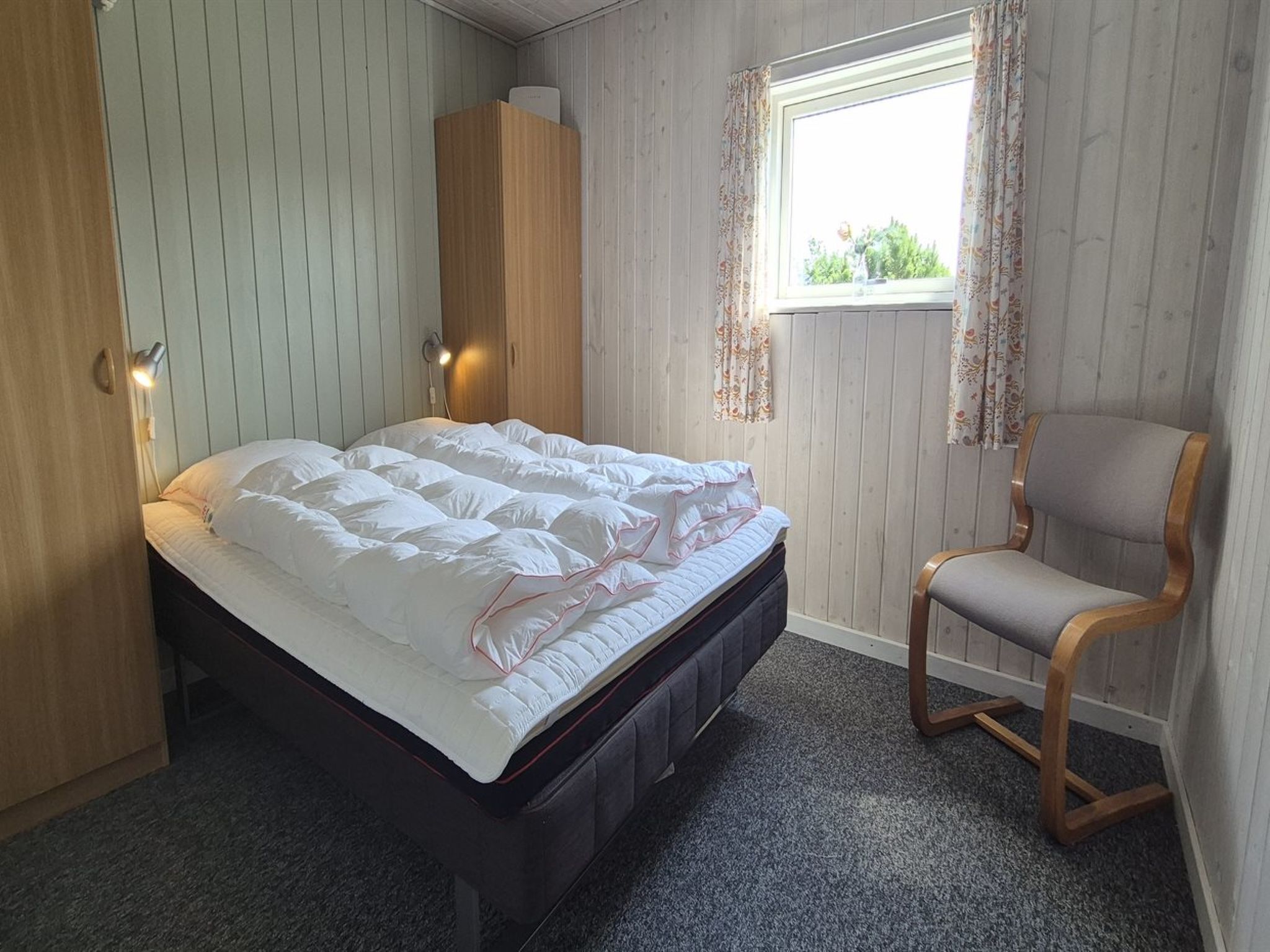 Un dormitorio con cama, una silla, armario y ventana con cortinas.