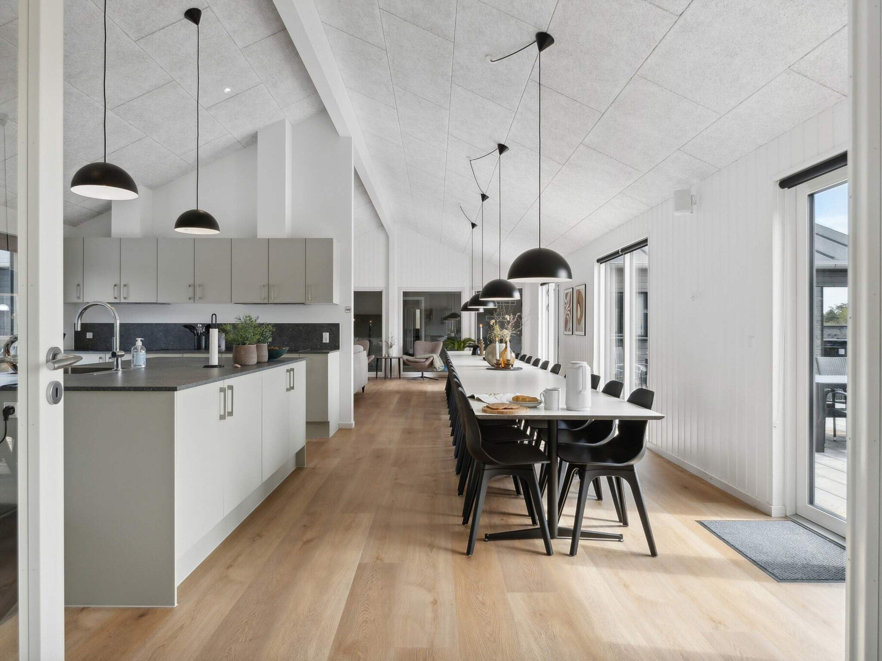 A modern kitchen with a central island, black pendant lighting, and a dining area with a long table and chairs.
