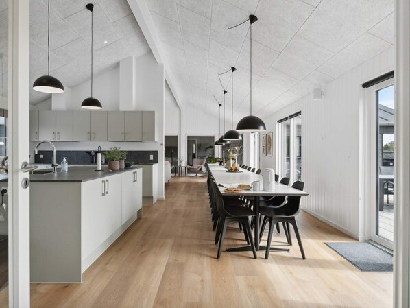 A modern kitchen with a central island, black pendant lighting, and a dining area with a long table and chairs. - 5 Star Holiday Home in Idestrup (Idestrup)