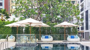 Zona de piscina con sillones de descanso y sombrillas, rodeada de vegetación y un edificio.