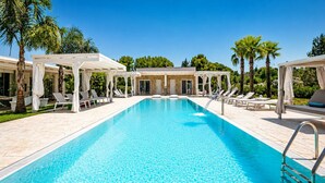 A rectangular swimming pool with a clear blue water, surrounded by white sun loungers and covered seating areas, with palm trees and a clear sky in the background. - Villa Rudiae by Symphonya Rentals (Lequile)