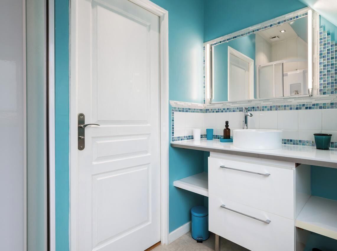 A bathroom with a white door, a sink with a mirror, and a blue trash bin.