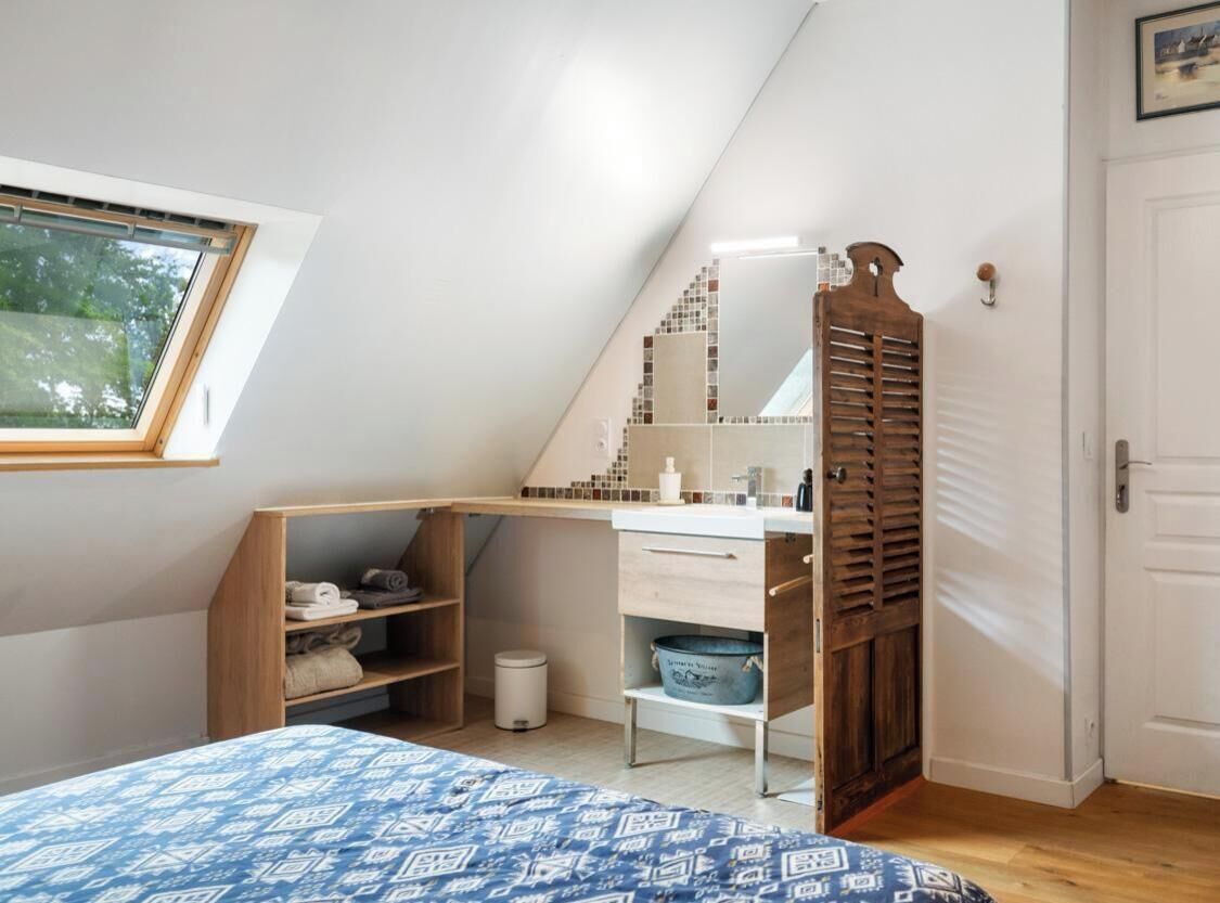 Une chambre avec un plafond en pente, un lit recouvert d’une couvre-lit à motifs, une armoire en bois, une salle de bain avec un mur recouvert de carreaux, et une fenêtre donnant sur des arbres.