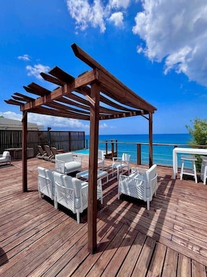 A wooden deck with white wicker furniture, a pergola, and a view of the ocean. - Villa Eve, 4-bedroom villa overlooking the ocean (Le Carbet)
