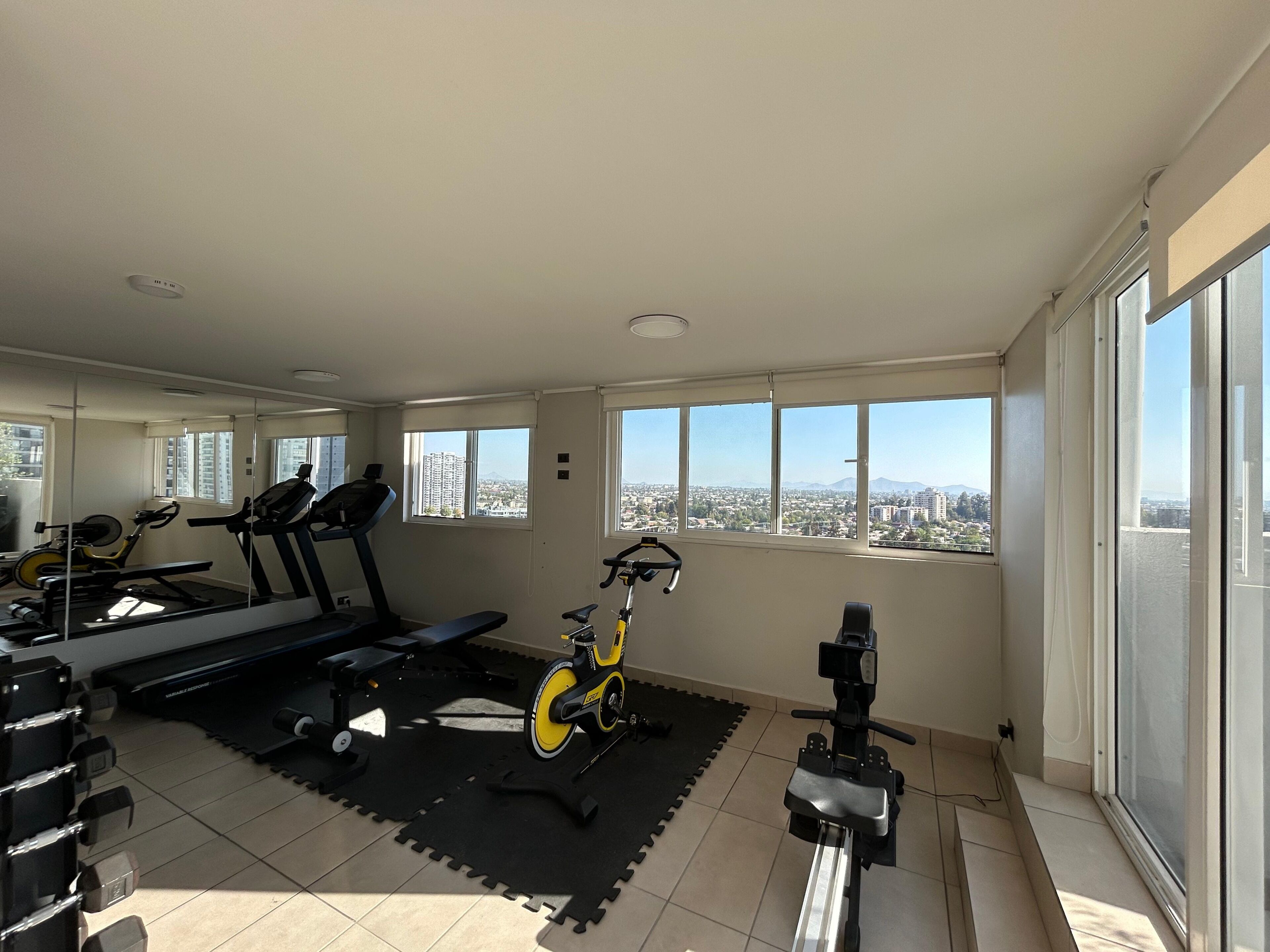 Un gimnasio espacioso con equipo de ejercicio, amplias ventanas y vista a la ciudad.