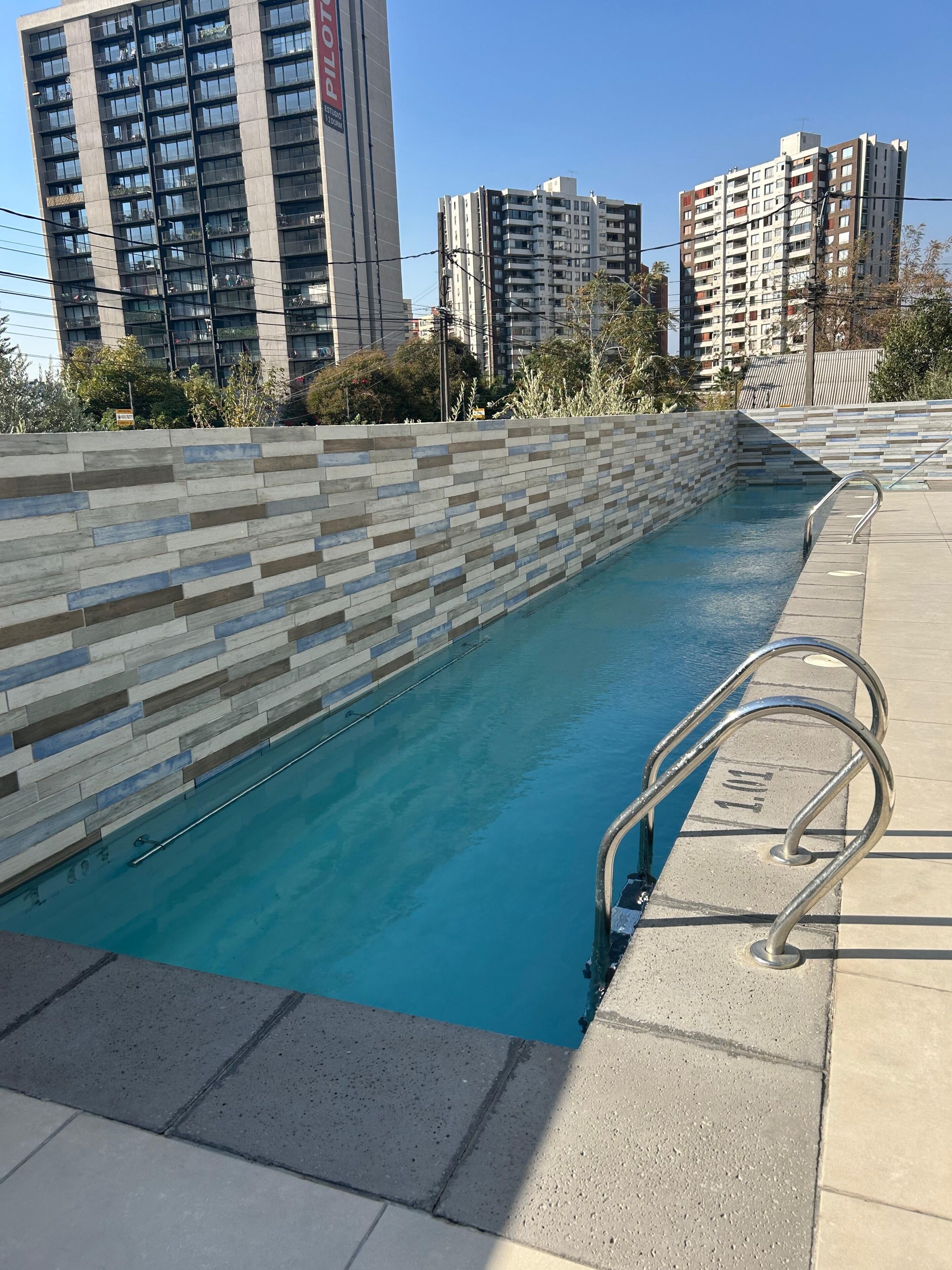 A swimming pool with a tiled wall and metal handrails, surrounded by high-rise buildings.