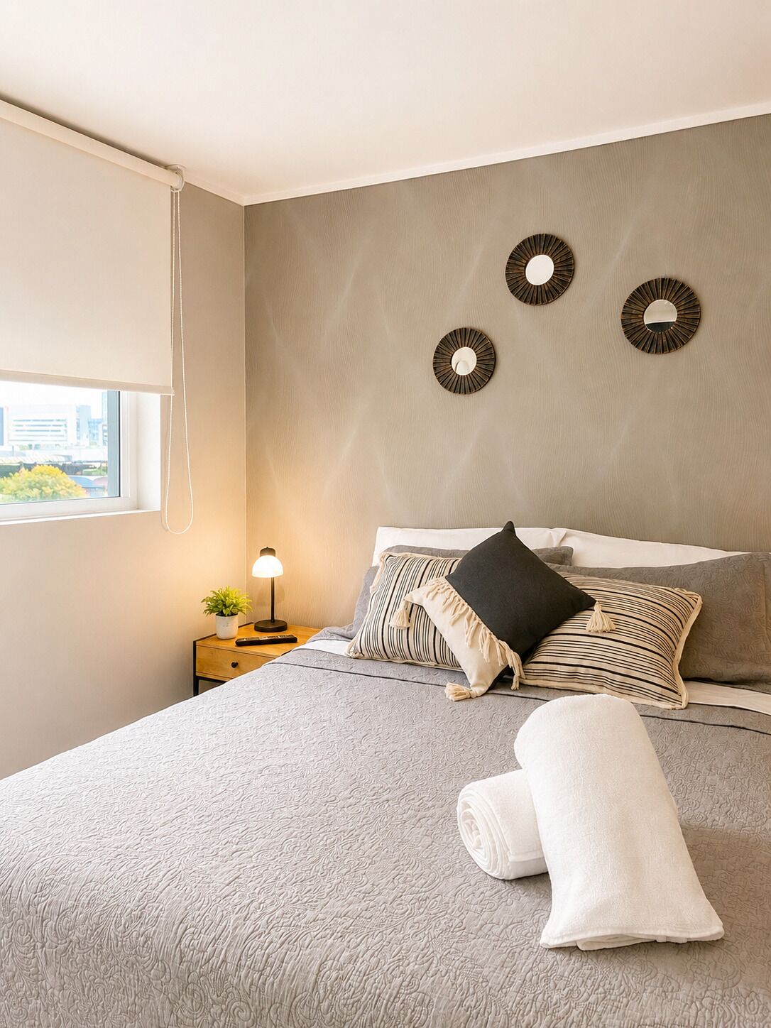 Una cama bien hecha con un edredón gris texturizado, una almohada negra y una almohada a rayas. Hay tres espejos circulares montados en la pared. Se ve una mesita de noche con una lámpara y una pequeña planta.