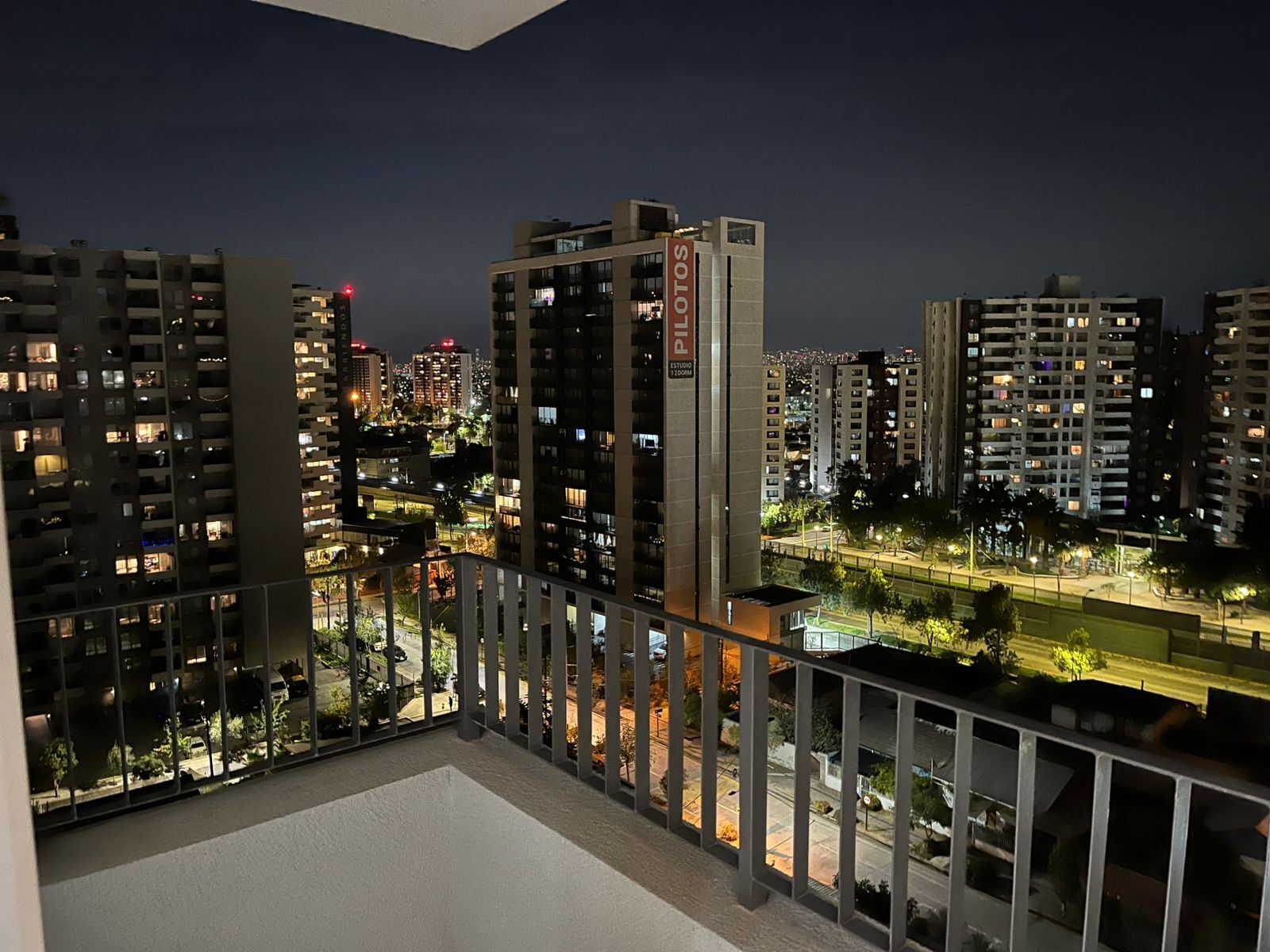 A nighttime view of a cityscape with high-rise buildings, a balcony with a railing, and a street below.