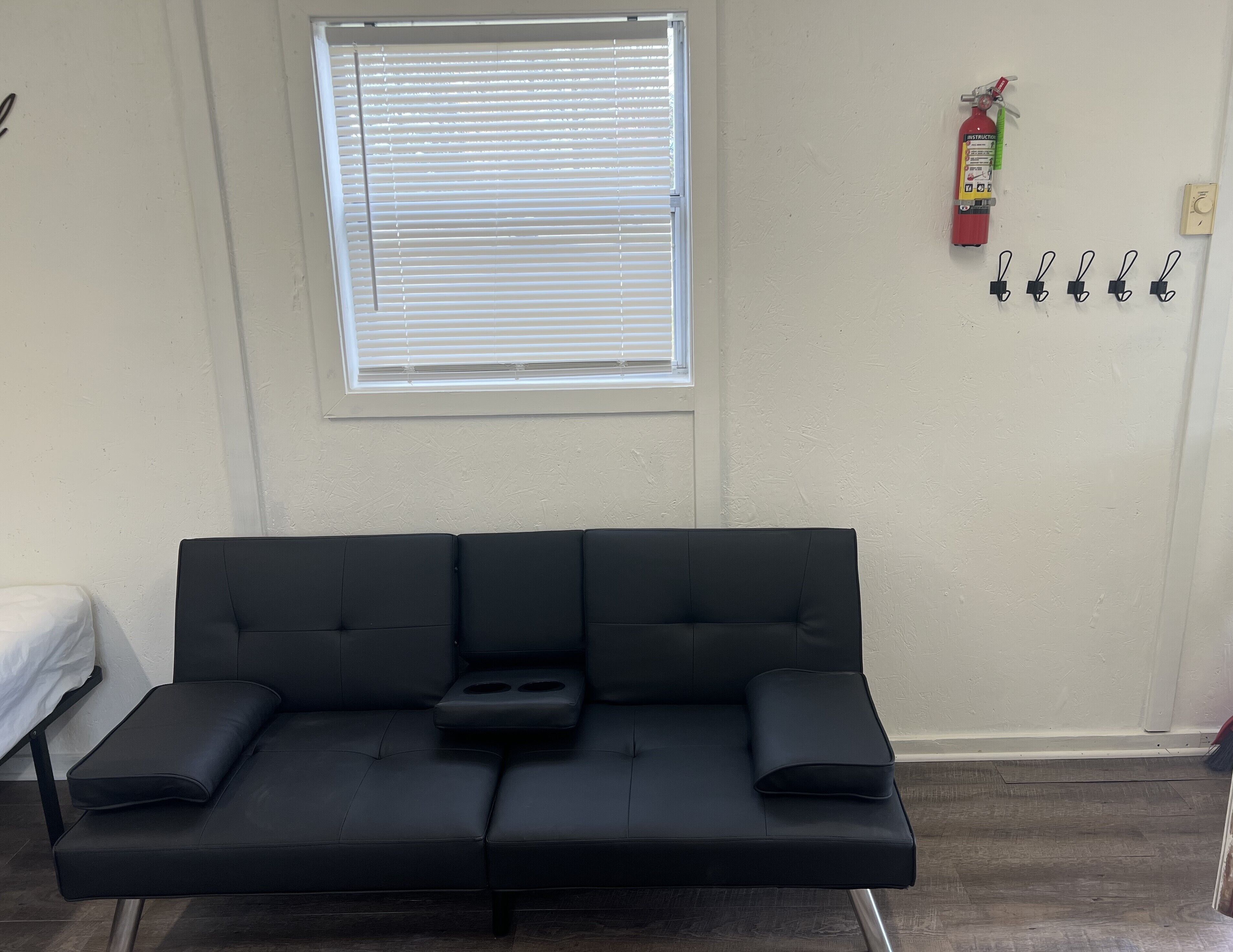 A black sofa with a tray table, a window with blinds, a fire extinguisher, and a wall-mounted light switch.